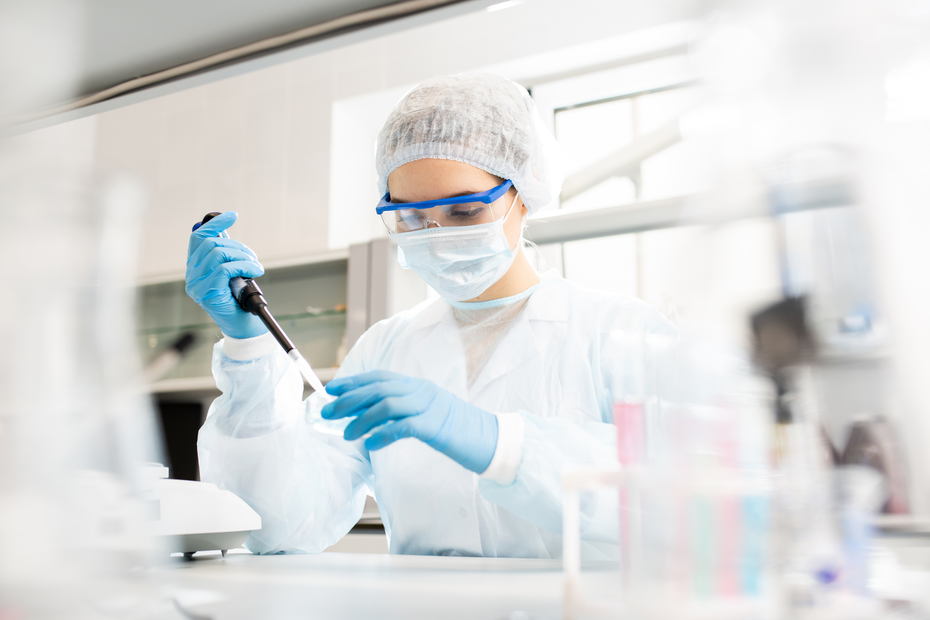 Laborantka w białym fartuchu i rękawicach, nosząca gogle i maseczkę, precyzyjnie używa pipety w laboratorium. W tle widoczny sprzęt laboratoryjny.