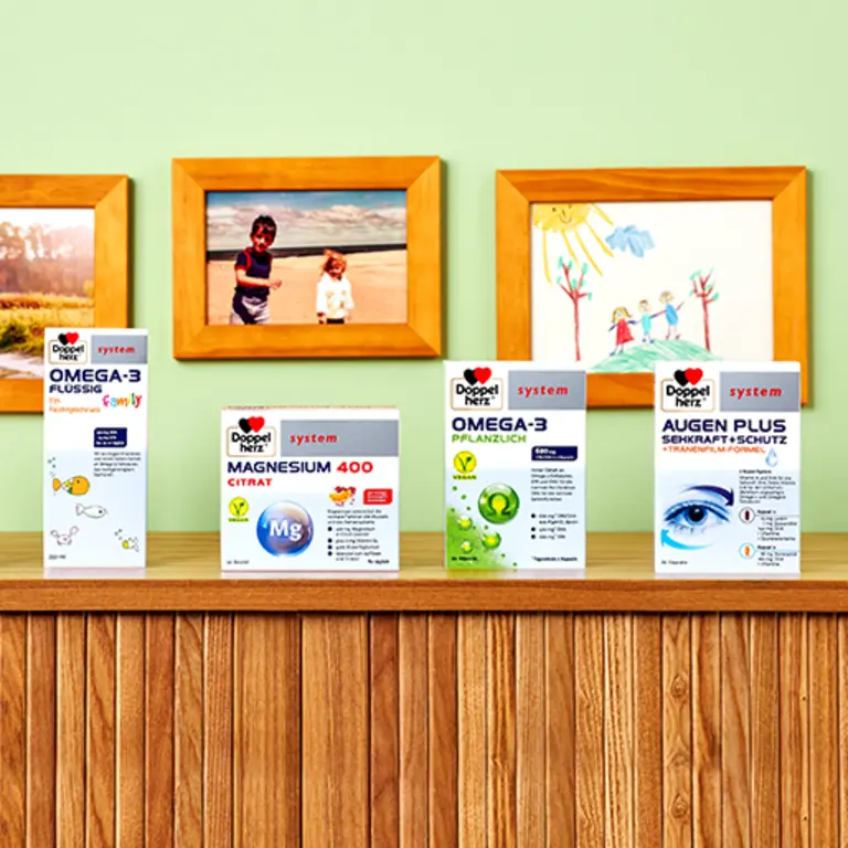 Na drewnianym stole znajdują się cztery produkty zdrowotne marki Doppelherz: OMEGA-3 płynny, MAGNEZ 400 CITRAT, OMEGA-3 roślinny oraz AUGEN PLUS z oznaczeniem ochrony wzroku. W tle widać kolorowe rysunki i zdjęcia na zielonej ścianie.
