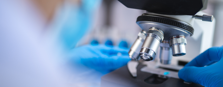 Mikroskop w laboratorium, na którym widoczne są ręce osoby w niebieskich rękawiczkach, ustawiającej próbkę. Tło jest rozmyte, a uwaga skupiona na mikroskopie i badaniu.