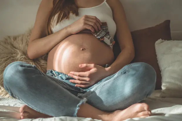 Kobieta siedzi z zaokrąglonym brzuchem, trzymając w dłoniach małe skarpetki dla niemowlęcia. Ma na sobie biały top i niebieskie dżinsy. Jej ręce spoczywają na brzuchu, co podkreśla jej ciążę. W tle widać miękkie tekstury i neutralne kolory.