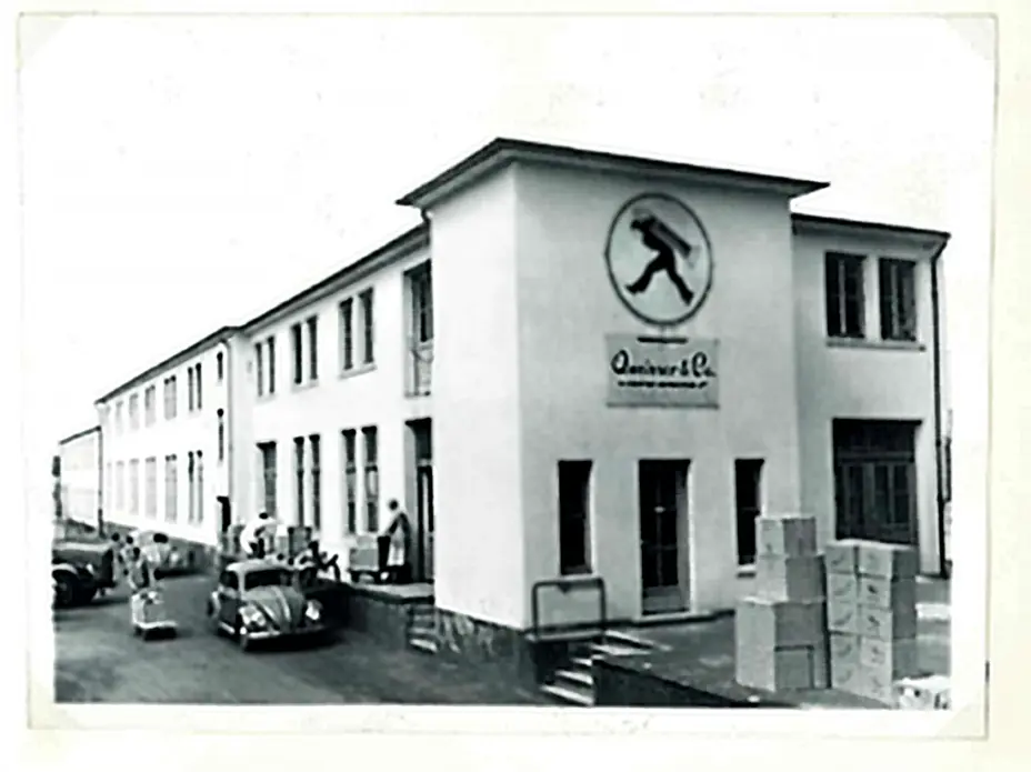Budynek firmy z logo na ścianie, w stylu retro. Przed nim stoją samochody oraz osoby zajęte pracą. W tle widać kartony ułożone na schodkach.