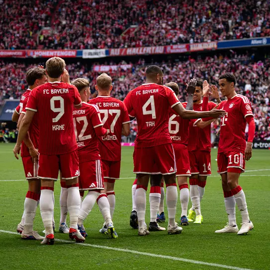 Zawodnicy FC Bayern Monachium w czerwonych strojach, z widocznymi numerami na koszulkach, zbierają się w grupie na boisku. W tle widać tłum kibiców. Atmosfera jest radosna, a gracze wydają się przygotowani do meczu.