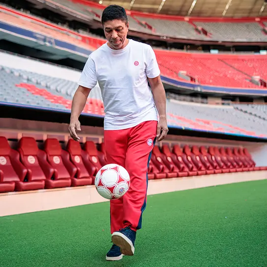 Mężczyzna w białej koszulce i czerwonych spodniach trenuje na stadionie piłkarskim, kopiąc piłkę. W tle widać puste trybuny. Atmosfera jest sportowa i dynamiczna.