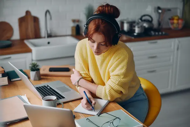 Młoda kobieta w żółtym swetrze, nosząca słuchawki, siedzi przy stole w kuchni. Pisze w notesie, obok niej laptop i kubek z napojem. W tle widać kuchenne akcesoria. Kobieta wydaje się skupiona i zorganizowana w swoim miejscu pracy.