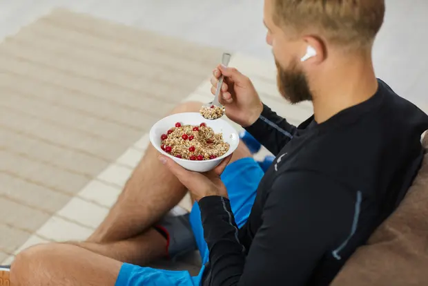 Mężczyzna w czarnym stroju sportowym siedzi na kanapie z misą pełną płatków śniadaniowych z czerwonymi owocami. Trzyma łyżkę w jednej ręce i zjeżdża z miski. W tle widoczny jest jasny dywan.